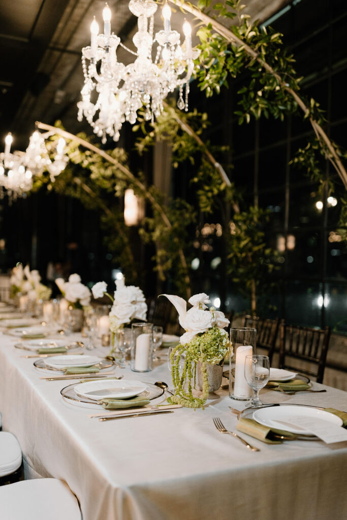 Indoor reception table under crystal chandeliers with greenery arches and candlelight.