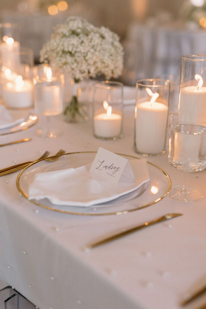Elegant wedding tablescape with neutral linens, layered place settings, and floral centerpieces in a softly lit reception space.