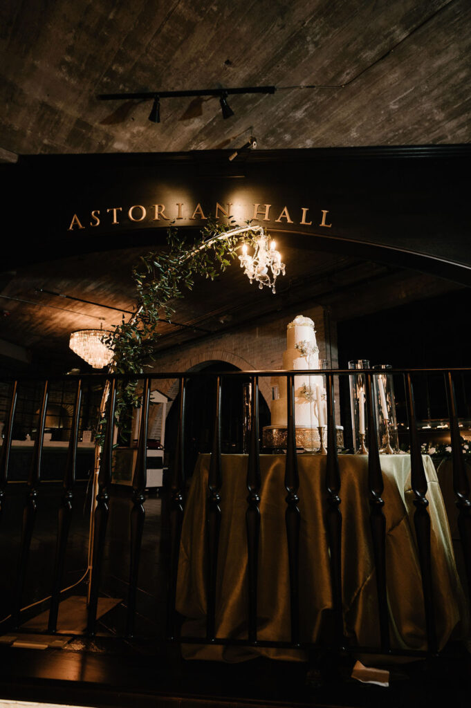 Wedding cake displayed at The Astorian Houston under chandelier lighting with greenery and “Astorian Hall” sign in the background